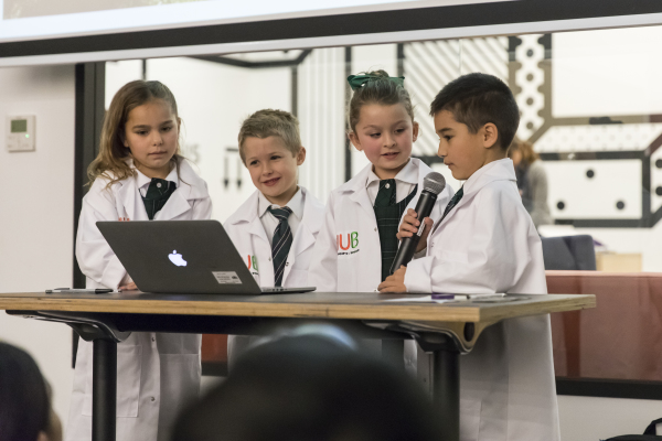 Group of children wearing white lab coats standing at a desk with a computer. One is speaking into a microphone.